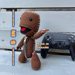 May include: A handmade brown crochet doll with a zipper down the front and white stitching. The doll has a black mouth and eyes, and a red tongue. A black game controller is next to the doll. The text "MADE IN UKRAINE" is visible.