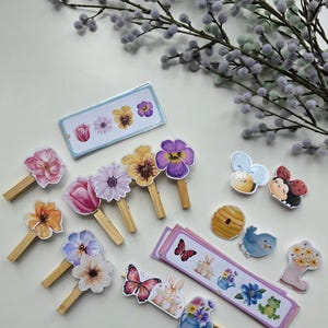 May include: A collection of floral and spring-themed decorations. Features flower-shaped pegs, stickers of butterflies, bees, bunnies, and a frog. A sprig of grey-green fuzzy buds is in the background. The items are arranged on a white surface.