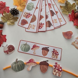Op de afbeelding: Een verzameling rechthoekige papieren bladwijzers en uitsparingen met herfstthema-illustraties van groene pompoenen, paddenstoelen met rode hoeden, bruine eikels en kleine bosdieren, gerangschikt met kunstmatige herfstbladeren.