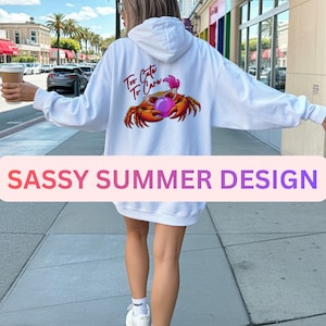 May include: White hoodie with a crab graphic wearing sunglasses and holding a pink bubble. The text "Too Cute To Care" is above the crab. The bottom of the image has the text "SASSY SUMMER DESIGN".