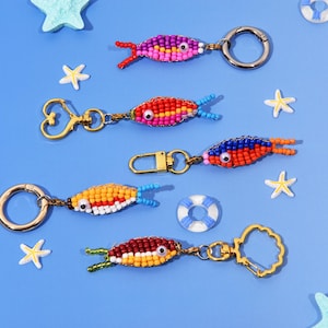 Puede incluir: Varios llaveros de peces hechos con cuentas de colores, incluyendo rosa, rojo, naranja y marrón, con detalles azules y verdes. Los llaveros tienen herrajes dorados o plateados y están colocados sobre un fondo azul con estrellas de mar y un salvavidas en miniatura.