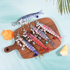 Może przedstawiać: Breloki w kształcie ryb w kolorach niebieskim, czerwonym i fioletowym, z detalami w postaci gwiazd i koralików. Breloki mają srebrne metalowe okucia i są prezentowane na drewnianej desce. Deska jest ozdobiona muszlami i gwiazdami.