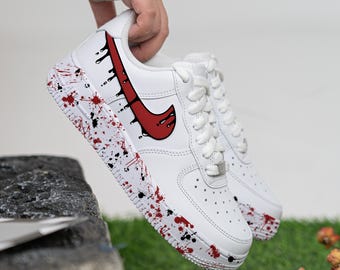 May include: White low-top trainers with a red and black paint splatter design. The Nike swoosh is red and has a dripping effect.