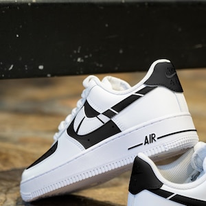 May include: White leather low-top sneakers with black accents. The Nike swoosh logo is outlined in black and the word "AIR" is printed on the side of the shoe.