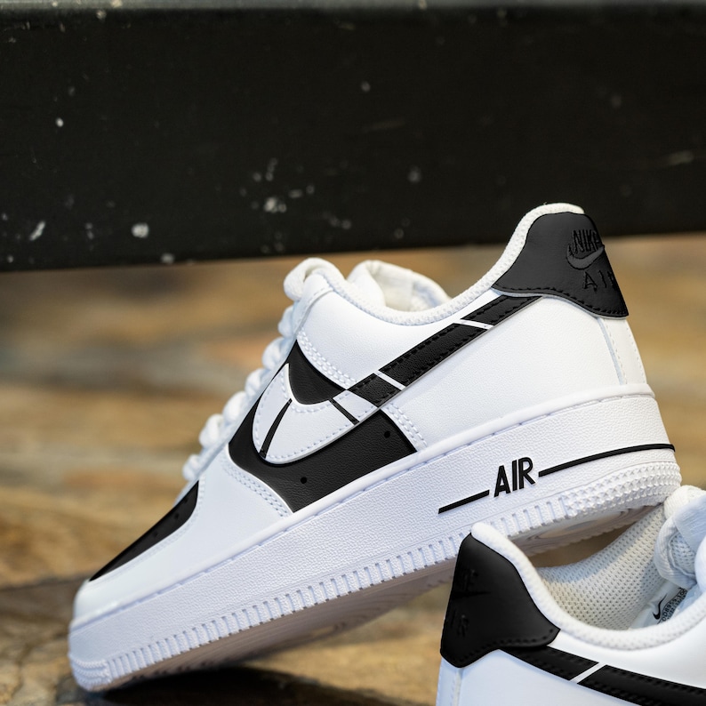 May include: White leather low-top sneakers with black accents. The Nike swoosh logo is outlined in black and the word "AIR" is printed on the side of the shoe.