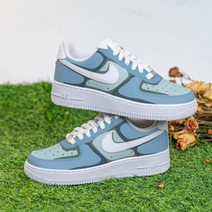 Puede incluir: Un par de zapatillas Nike Air Force 1 azules y blancas con un dise&ntilde;o abstracto &uacute;nico. Las zapatillas tienen una suela blanca y una parte superior azul con detalles blancos.