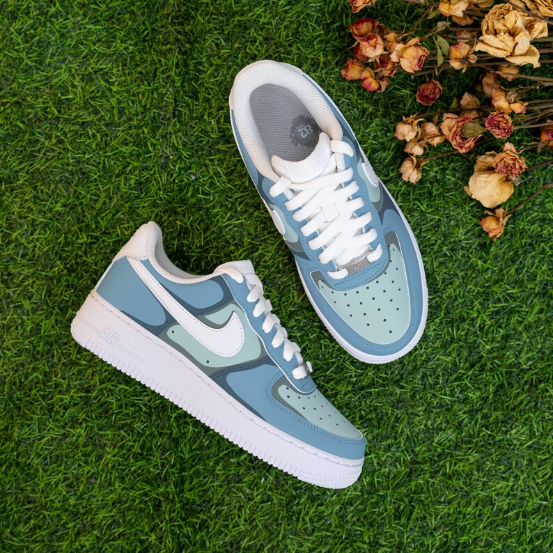 Puede incluir: Un par de zapatillas Nike Air Force 1 azules y blancas con un patr&oacute;n de camuflaje. Las zapatillas tienen cordones blancos y una suela blanca.