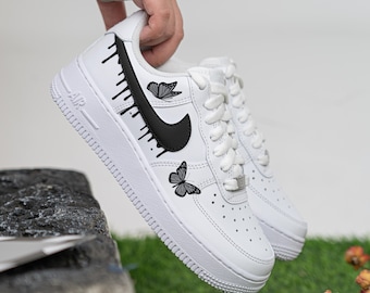 May include: White leather low-top sneakers with a black swoosh and black butterfly designs. The sneakers have a white rubber sole and a black drip design on the side.