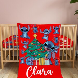 May include: Red and green Christmas themed blanket with a blue Stitch character from Lilo & Stitch holding a Christmas tree. The blanket has a white name "Clara" on the bottom.