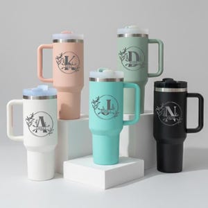 May include: A collection of personalized insulated tumblers in various colors, including white, pink, mint green, and black. Each tumbler features a handle, a lid, and a custom initial and name design. The tumblers are designed for hot or cold beverages.