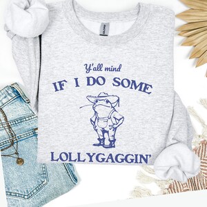 May include: A light gray sweatshirt with the text "Y'all mind IF I DO SOME LOLLYGAGGIN'" and a blue illustration of a frog wearing a cowboy hat and overalls. The sweatshirt is paired with denim shorts and a necklace.