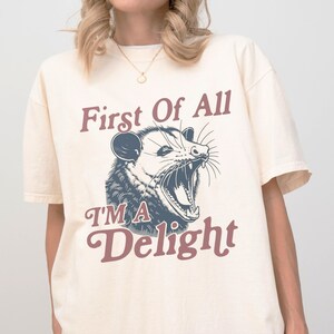 May include: Cream-colored t-shirt featuring a graphic of an opossum with an open mouth. The text on the shirt reads "First Of All I'M A Delight" in maroon and dark blue lettering. The shirt has short sleeves and a relaxed fit.
