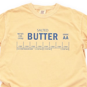 May include: Butter-colored t-shirt with the word "BUTTER" in large blue letters. The shirt also features text indicating net weight, grade, and measurement markings for butter, including "4 OZ. (113g)" and "1/2 CUP".