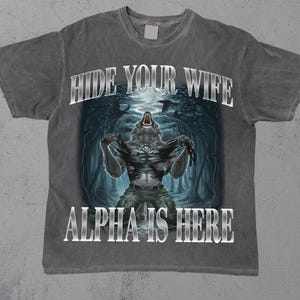 Puede incluir: Una camiseta gris con un aspecto desgastado presenta un gráfico de un hombre lobo con el texto "Hide Your Wife Alpha Is Here".