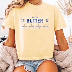 Puede incluir: Una camiseta amarilla Comfort Colors boxy/cropped con la palabra "BUTTER" en letras azules. La camiseta tiene un gr&aacute;fico de medidas de mantequilla, incluyendo "NET WT. 4 oz. (113g)" y "GRADE AA".