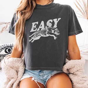 May include: Dark grey boxy crop tee featuring the word "EASY" above a white tiger graphic. The shirt is a comfort colour and has a crew neck. The model is wearing denim shorts and a cream-coloured sweater.