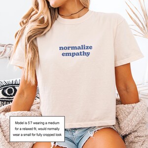May include: Cream-colored t-shirt with the words "normalize empathy" in blue lettering. The shirt has short sleeves and a relaxed fit. The model is wearing a medium for a relaxed fit, or a small for a cropped look.