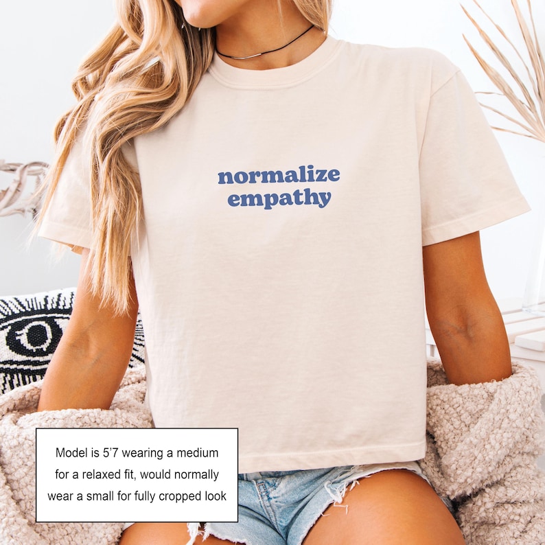 May include: Cream-colored t-shirt with the words "normalize empathy" in blue lettering. The shirt has short sleeves and a relaxed fit. The model is wearing a medium for a relaxed fit, or a small for a cropped look.
