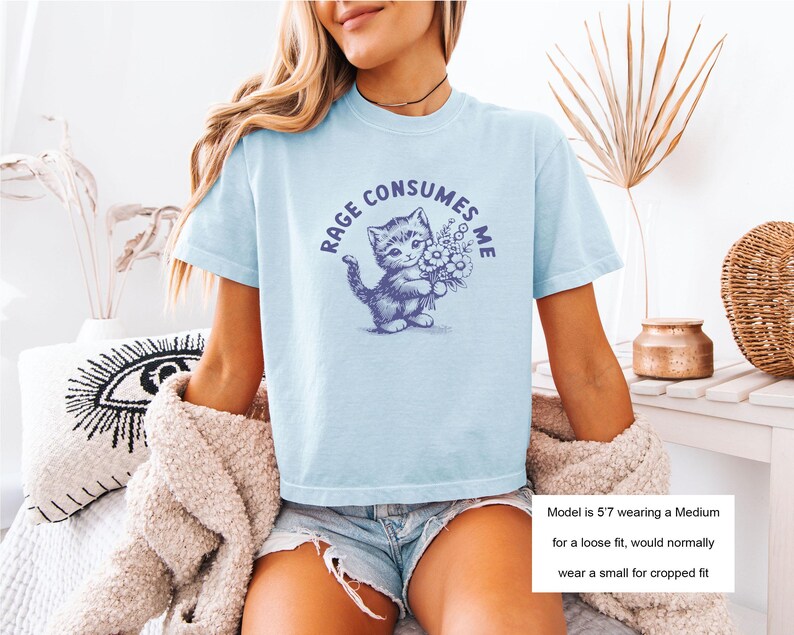 May include: Light blue t-shirt with a graphic of a kitten holding flowers and the text "RAGE CONSUMES ME" in a circular design. The shirt is a light blue color.