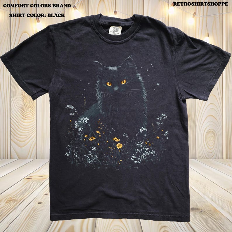 May include: Black t-shirt featuring a detailed illustration of a black cat with yellow eyes, surrounded by flowers and foliage. The shirt has the text "COMFORT COLORS BRAND" and "SHIRT COLOR: BLACK" at the top. The shop name "RETROSHIRTSHOPPE" is also visible.