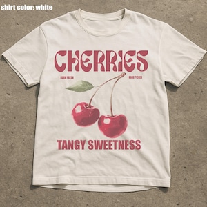 May include: White t-shirt with a red graphic of two cherries and the word "CHERRIES" in a stylized font. The text "FARM FRESH" and "HAND PICKED" are above the cherries, and "TANGY SWEETNESS" is below.