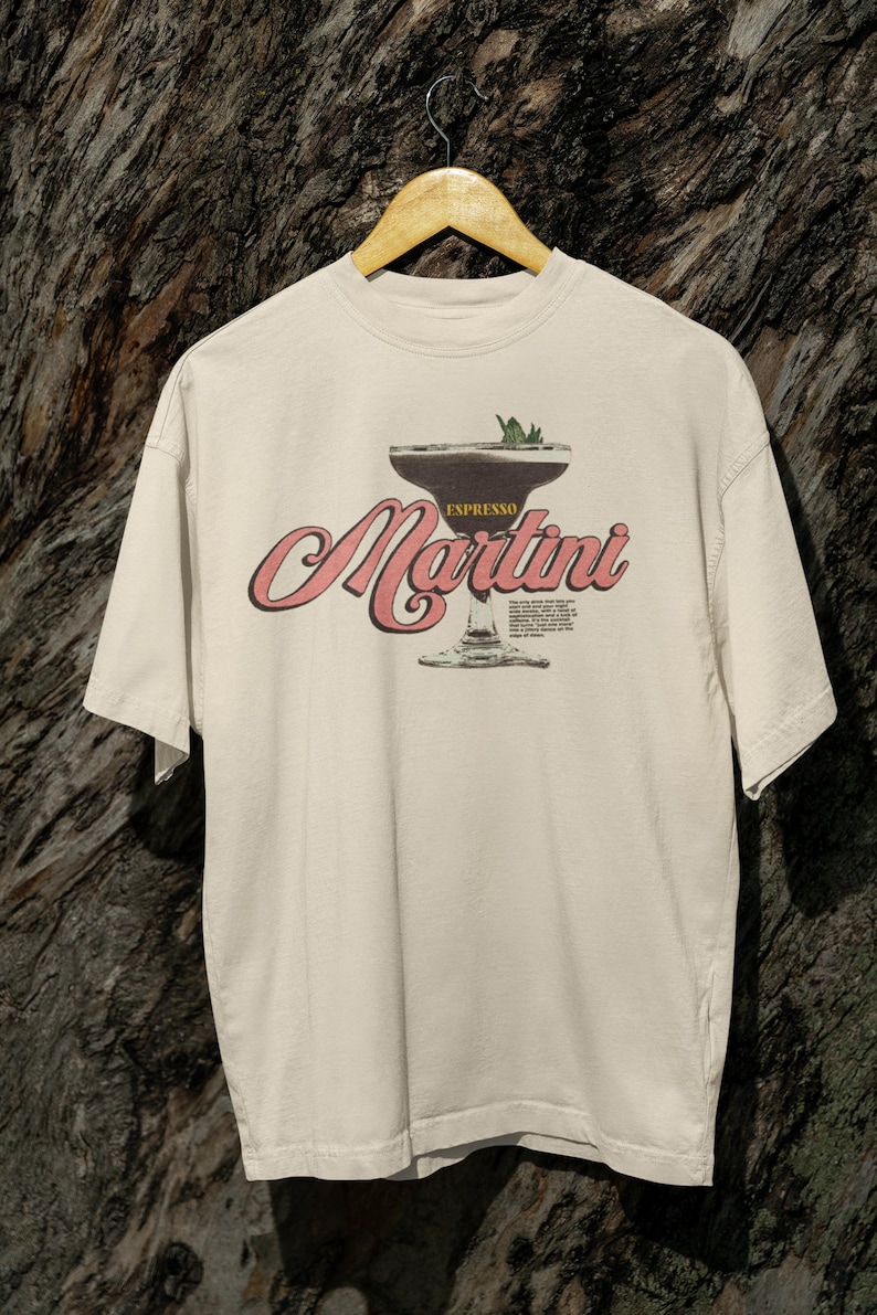 Puede incluir: Camiseta color crema con un gr&aacute;fico de un espresso martini. El dise&ntilde;o incluye la palabra "Espresso" encima de la copa de martini y la palabra "Martini" en cursiva. La camiseta est&aacute; colgada en una percha de madera.