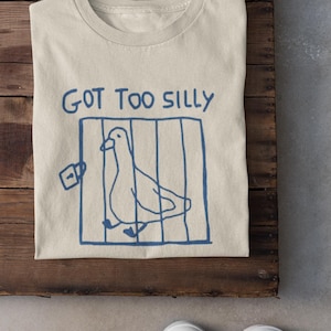May include: A beige t-shirt featuring a blue drawing of a pigeon in a cage and the words "GOT TOO SILLY". The shirt is folded on a wooden surface, with white trainers in the foreground.