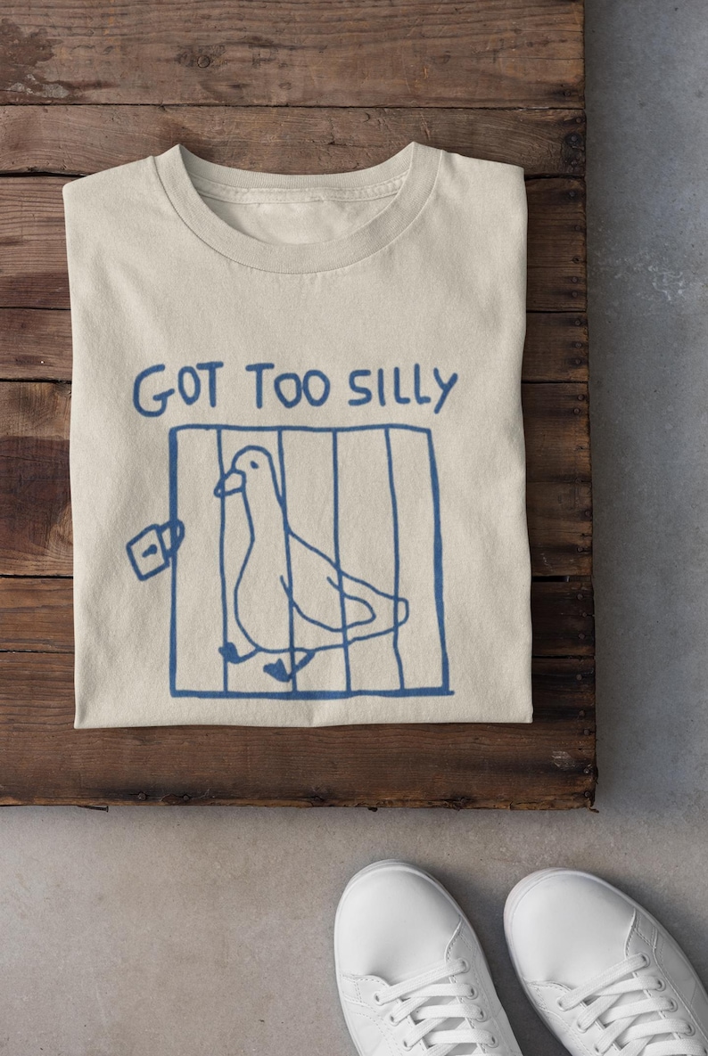 May include: A beige t-shirt featuring a blue drawing of a pigeon in a cage and the words "GOT TOO SILLY". The shirt is folded on a wooden surface, with white trainers in the foreground.