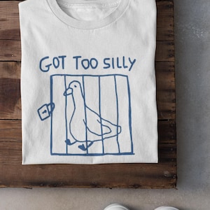 May include: White t-shirt featuring a blue drawing of a bird in a cage with the words "GOT TOO SILLY". The shirt is on a wooden surface, with white trainers visible in the foreground.