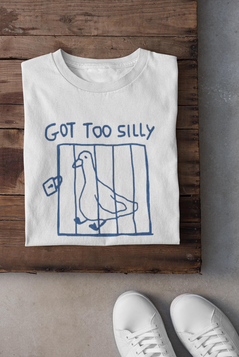 May include: White t-shirt featuring a blue drawing of a bird in a cage with the words "GOT TOO SILLY". The shirt is on a wooden surface, with white trainers visible in the foreground.