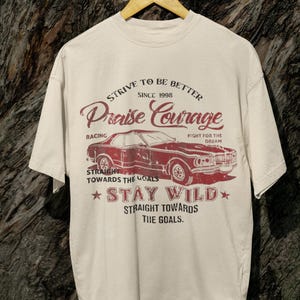K&ouml;nnte beinhalten: Ein beiges T-Shirt mit einem roten Vintage-Auto-Motiv und dem Text "Strive to be better since 1998 Praise Courage Racing Fight for the Dream Straight towards the goals Stay Wild Straight towards the goals."