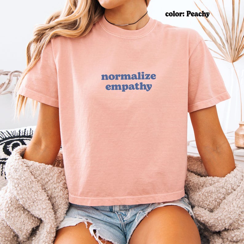 May include: A peach-colored t-shirt with the words "normalize empathy" in blue lettering. The shirt is short-sleeved and has a crew neck. The model is wearing denim shorts and a necklace.