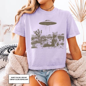 May include: Orchid-colored comfort colors cropped shirt featuring a graphic of a cat being beamed up by a UFO in a desert landscape. The shirt has short sleeves and a crew neck. The text on the bottom of the shirt reads: SHIRT COLOR: ORCHID COMFORT COLORS CROPPED SHIRT.