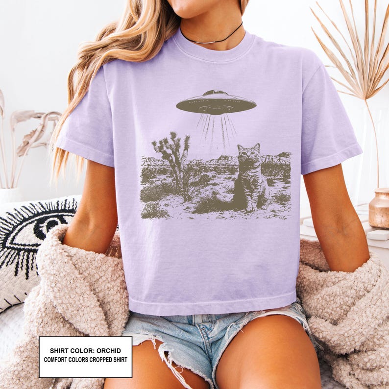 May include: Orchid-colored comfort colors cropped shirt featuring a graphic of a cat being beamed up by a UFO in a desert landscape. The shirt has short sleeves and a crew neck. The text on the bottom of the shirt reads: SHIRT COLOR: ORCHID COMFORT COLORS CROPPED SHIRT.