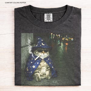 Peut inclure: T-shirt gris fonc&eacute; avec un motif de chat portant un chapeau de sorcier, des lunettes de soleil et une cape, fumant une cigarette. Le chat est assis sur un trottoir avec un arri&egrave;re-plan flou. Le t-shirt porte le texte "COMFORT COLORS PEPPER".