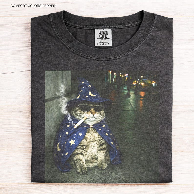 Peut inclure: T-shirt gris fonc&eacute; avec un motif de chat portant un chapeau de sorcier, des lunettes de soleil et une cape, fumant une cigarette. Le chat est assis sur un trottoir avec un arri&egrave;re-plan flou. Le t-shirt porte le texte "COMFORT COLORS PEPPER".