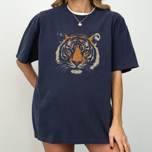 K&ouml;nnte beinhalten: Marineblaues T-Shirt mit einem Tigergesicht-Motiv in Orange und Beige. Das Design ist mittig auf der Vorderseite. Das T-Shirt hat kurze &Auml;rmel und eine lockere Passform. Die Person tr&auml;gt eine goldene Halskette.