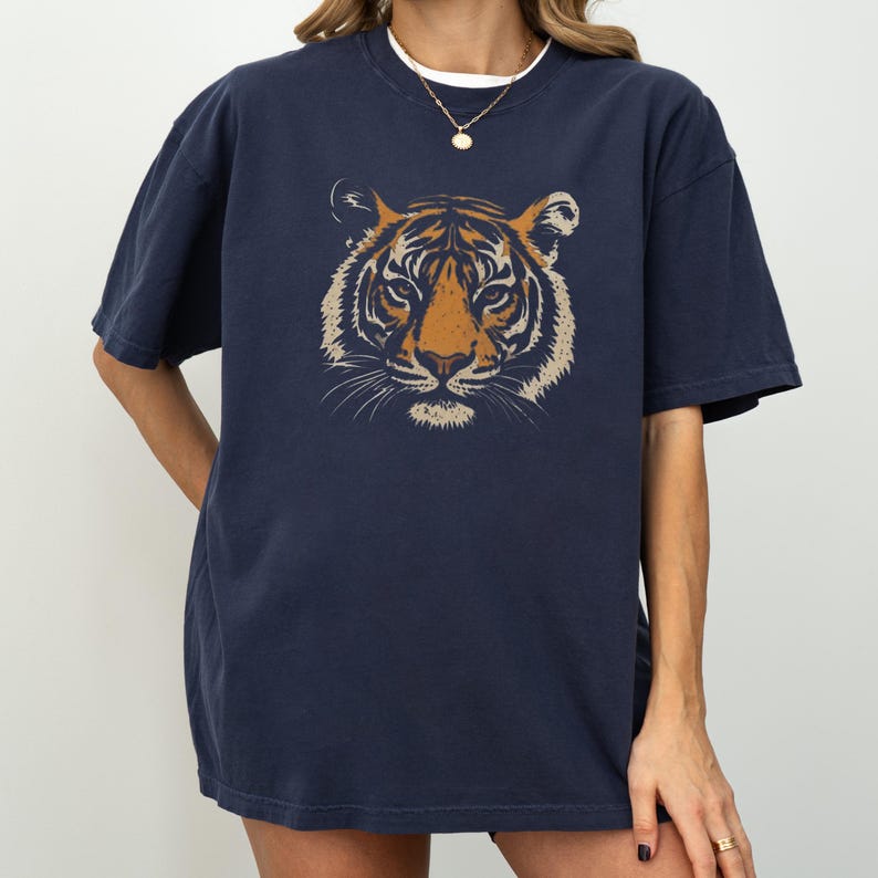 K&ouml;nnte beinhalten: Marineblaues T-Shirt mit einem Tigergesicht-Motiv in Orange und Beige. Das Design ist mittig auf der Vorderseite. Das T-Shirt hat kurze &Auml;rmel und eine lockere Passform. Die Person tr&auml;gt eine goldene Halskette.
