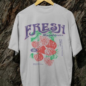 May include: A white t-shirt with a graphic print of raspberries and the text "FRESH Raspberries Enjoy Little Things While They Last Sweet Fresh And Fruity".