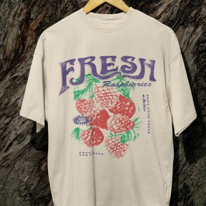May include: A beige t-shirt with a graphic print of raspberries and the text "FRESH Raspberries Enjoy Little Things Sweet Fresh And Tart".