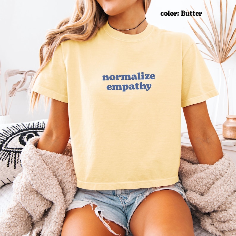 May include: Butter-colored t-shirt with the words "normalize empathy" in blue. The shirt is short-sleeved and appears to be made of a soft material. The model is wearing denim shorts and a beige sweater.