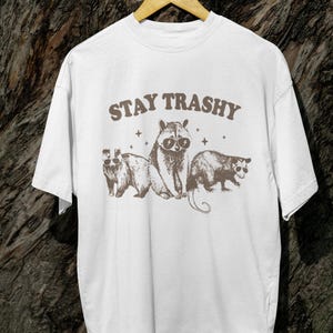K&ouml;nnte beinhalten: Wei&szlig;es T-Shirt mit dem Text "STAY TRASHY" in braunen Buchstaben. Das Shirt zeigt eine Grafik von drei Tieren mit Sonnenbrillen: einem Waschb&auml;ren, einem Opossum und einem Dachs.