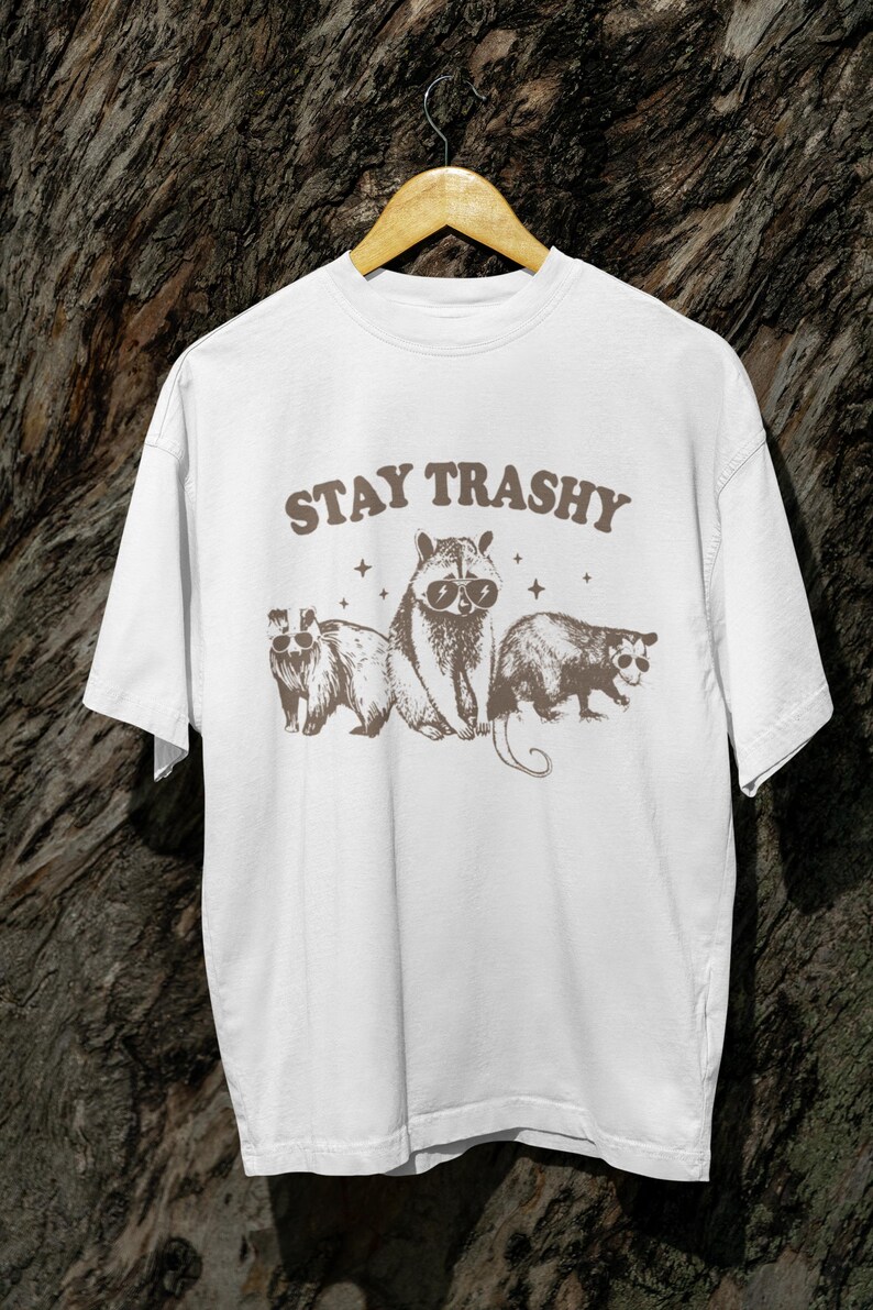 K&ouml;nnte beinhalten: Wei&szlig;es T-Shirt mit dem Text "STAY TRASHY" in braunen Buchstaben. Das Shirt zeigt eine Grafik von drei Tieren mit Sonnenbrillen: einem Waschb&auml;ren, einem Opossum und einem Dachs.