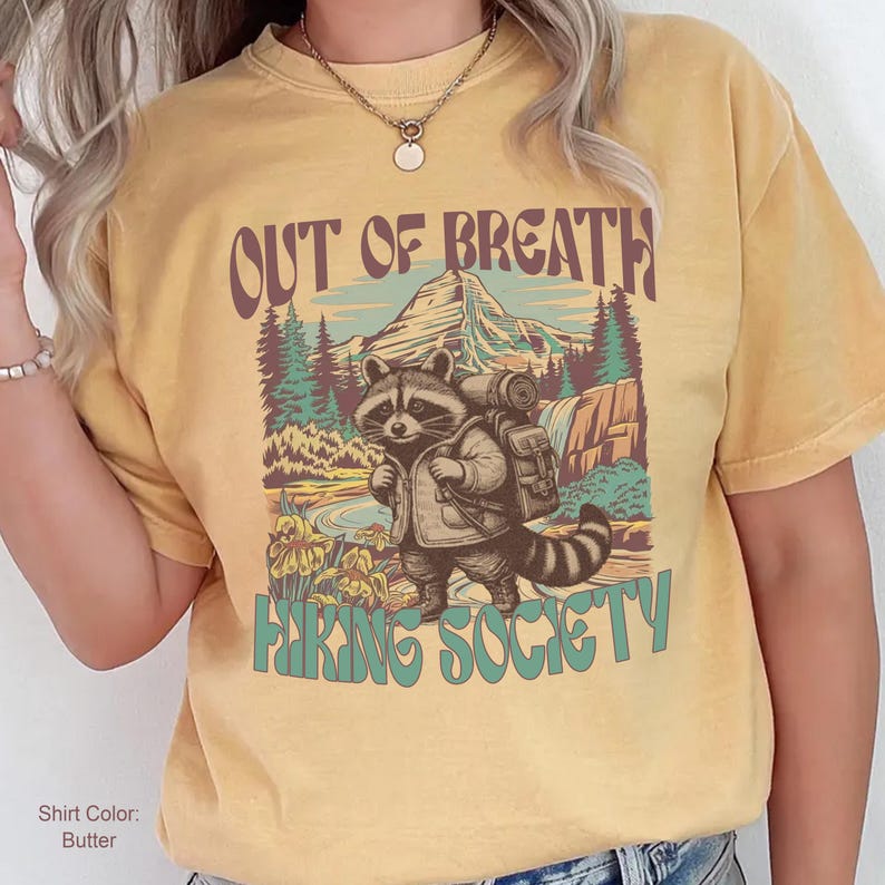 May include: Butter-colored t-shirt with a graphic of a raccoon wearing a backpack, standing in front of a mountain and waterfall scene. Text on the shirt reads "OUT OF BREATH" and "HIKING SOCIETY" in brown and teal lettering.