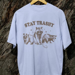 K&ouml;nnte beinhalten: Ein hellblaues T-Shirt mit einer braunen Grafik eines Waschb&auml;ren, einer Opossum und eines Opossums mit Sonnenbrille. Der Text "STAY TRASHY" ist in Braun &uuml;ber den Tieren gedruckt.