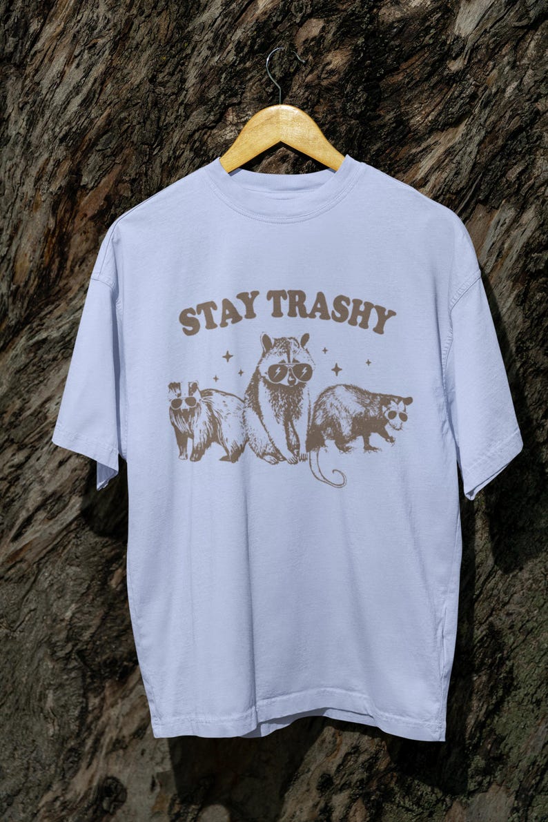 K&ouml;nnte beinhalten: Ein hellblaues T-Shirt mit einer braunen Grafik eines Waschb&auml;ren, einer Opossum und eines Opossums mit Sonnenbrille. Der Text "STAY TRASHY" ist in Braun &uuml;ber den Tieren gedruckt.