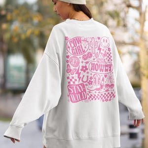 May include: White sweatshirt with a pink graphic design on the back. The design includes the words "Cow Girl", "Howdy", and "Stay Wild", along with various cowboy-themed illustrations such as boots, a cactus, and a horseshoe.