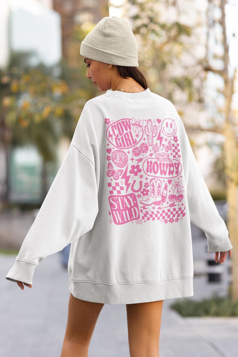 May include: White sweatshirt with a pink graphic design on the back. The design includes the words "Cow Girl", "Howdy", and "Stay Wild", along with various cowboy-themed illustrations such as boots, a cactus, and a horseshoe.