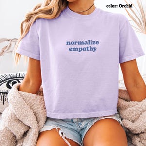 May include: Orchid-colored t-shirt with the words "normalize empathy" in blue lettering. The shirt has a crew neck and short sleeves. The model is wearing denim shorts and a beige sweater.