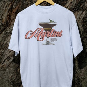 Puede incluir: Camiseta azul claro con un gr&aacute;fico de un espresso martini. El dise&ntilde;o incluye la palabra "Espresso" encima de la copa de martini y la palabra "Martini" en cursiva. La camiseta est&aacute; colgada en una percha de madera sobre un fondo texturizado.